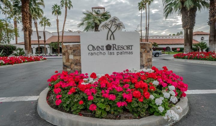 Entrance sign of Omni Rancho Las Palmas Resort, surrounded by red and pink flowers, with palm trees and a cloudy sky in the background.