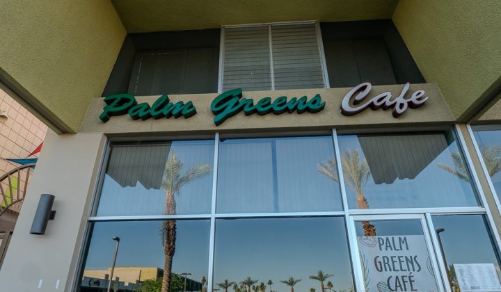 Exterior view of Palm Greens Café, featuring large storefront windows, palm trees reflected on the glass, and green and pink signage above the entrance.