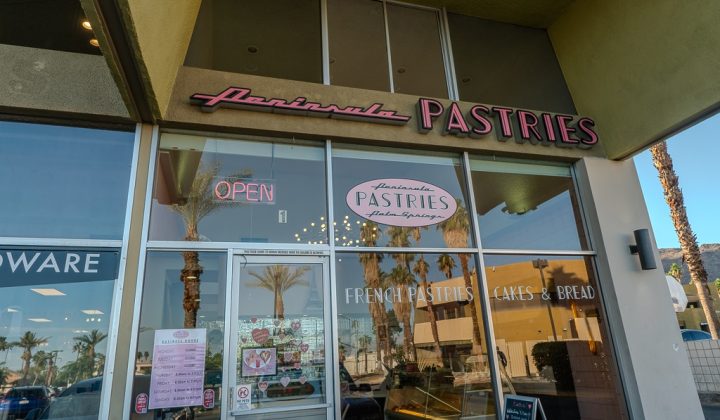 Storefront of Peninsula Pastries with glass windows, reflecting palm trees. Sign reads "French Pastries, Cakes & Bread." An "OPEN" sign is lit, and a menu is posted on the door.