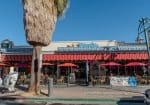 A cafe with red umbrellas and outdoor seating, near a large palm tree. A statue stands in front. Signage reads "Pinocchio." Clear sky in the background.