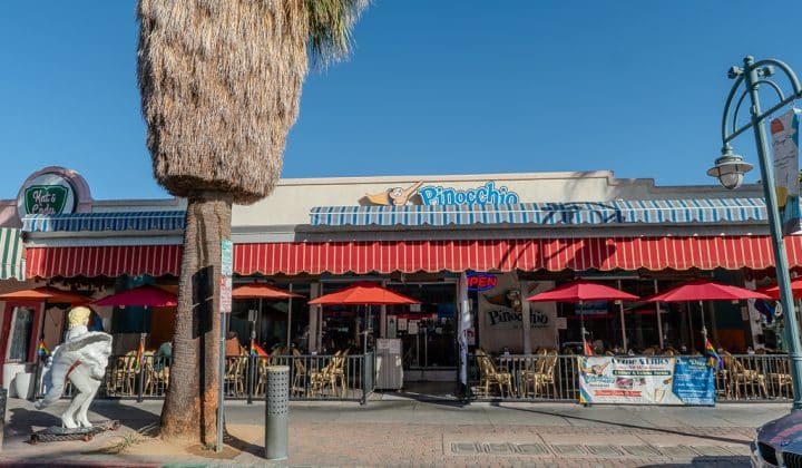 A cafe with red umbrellas and outdoor seating, near a large palm tree. A statue stands in front. Signage reads "Pinocchio." Clear sky in the background.