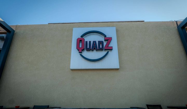 Sign with the text "QUADZ" on a beige wall above outdoor tables and chairs.