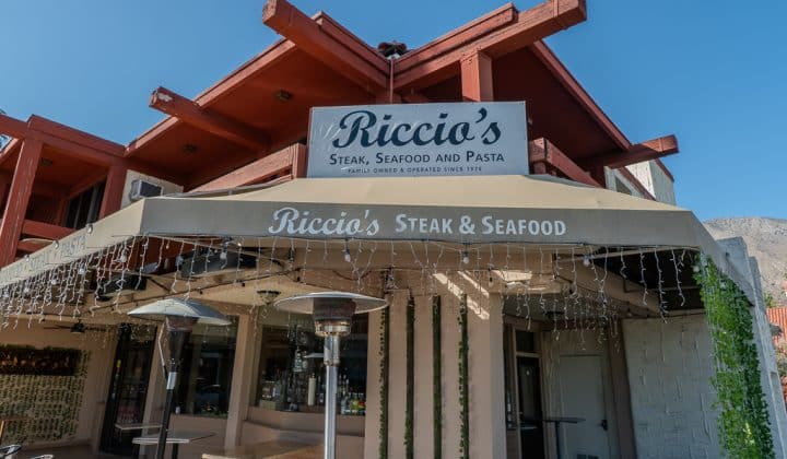 Exterior of Riccio's restaurant with signage for steak, seafood, and pasta under a red angular roof. White fairy lights hang from the awning.
