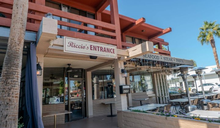 Exterior view of a restaurant named Riccio's with outdoor seating, a sign for seafood, steak, and pasta, and palm trees nearby.