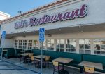 Outdoor seating area of a restaurant named "Rick's" with a tan stucco exterior and maroon signage. Tables and chairs are arranged near a handicapped parking sign.