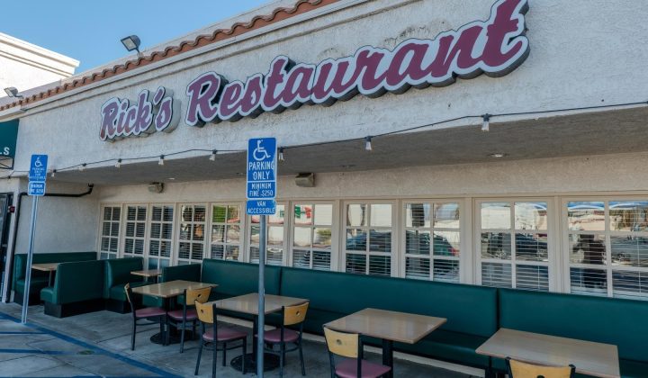 Outdoor seating area of a restaurant named "Rick's" with a tan stucco exterior and maroon signage. Tables and chairs are arranged near a handicapped parking sign.