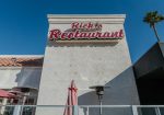 Exterior of "Rick's Restaurant" with a red and white sign. Umbrellas and patio heaters are visible, with a clear blue sky in the background.