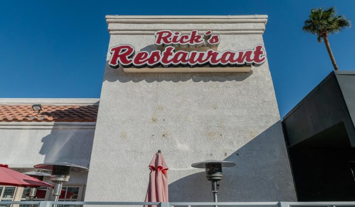 Exterior of "Rick's Restaurant" with a red and white sign. Umbrellas and patio heaters are visible, with a clear blue sky in the background.