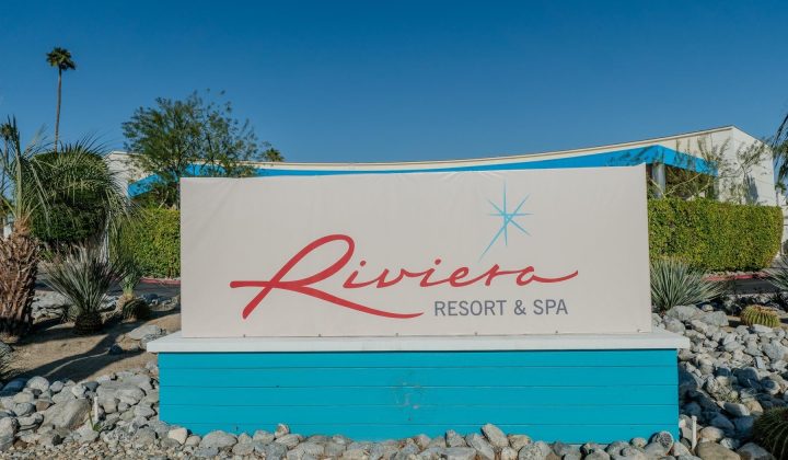 Sign reading "Riviera Resort & Spa" in red and blue text, surrounded by rocks and plants, with a clear blue sky in the background.