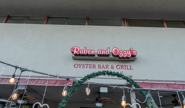 Sign for "Ruben and Ozzy's Oyster Bar & Grill" on a building. There's greenery and string lights hanging below.