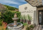 Entrance of Santiago with a fountain, potted plants, and a pergola. The building has a cream brick exterior with "Santiago" on the wall. Lush greenery is present in the surroundings.