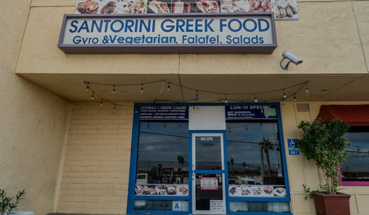 Front view of "Santorini Greek Food" restaurant with signage for gyro, vegetarian, falafel, and salads. Tan building, glass door, and string lights.