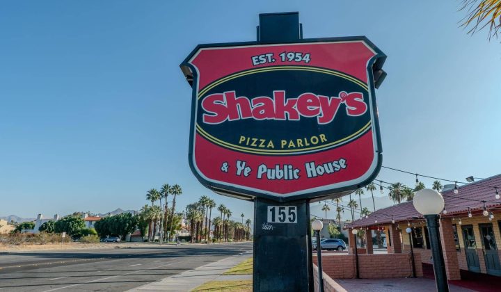 Sign for Shakey's Pizza Parlor, established 1954, with "Ye Public House" text, situated next to a road and palm trees.