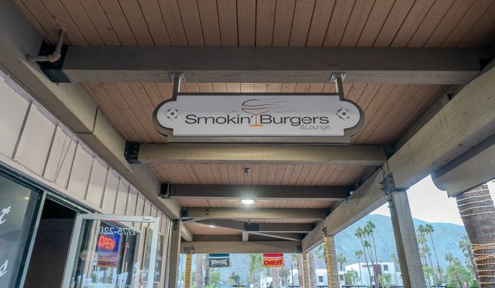 Sign for "Smokin' Burgers & Lounge" hangs under wooden ceiling of outdoor walkway, with view of a street and palm trees in the background.