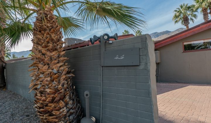 A brick wall with a mounted binocular viewfinder stands beside a palm tree, with mountains in the background.