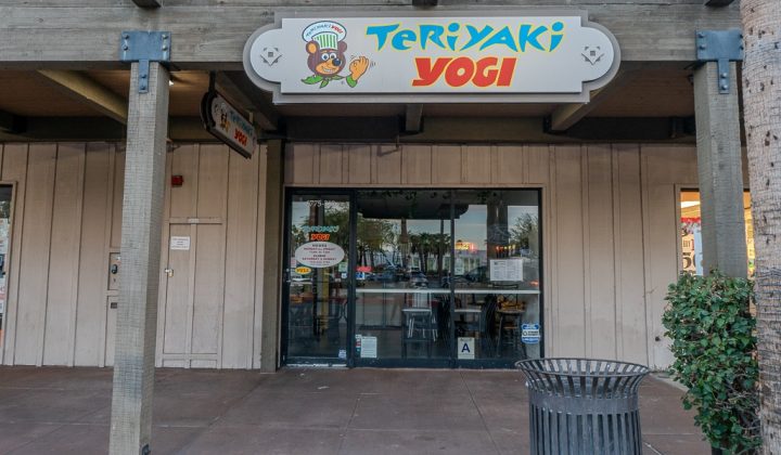 Front view of "Teriyaki Yogi" restaurant with a glass door and colorful sign. Wooden exterior with a trash can in the foreground. Reflections of cars and trees are visible in the glass.