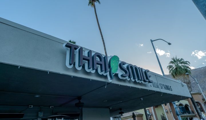 Sign for a restaurant named "Thai Smile" with palm trees and buildings in the background.