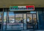 Front view of "The Real Italian Deli" with a glass door and window reflecting the outdoor surroundings. A mountain view is visible in the background.