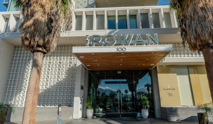 Entrance to a modern building with the name "Rowan" above the doors. Palm trees flank the entrance, and the building features large windows and a patterned facade.