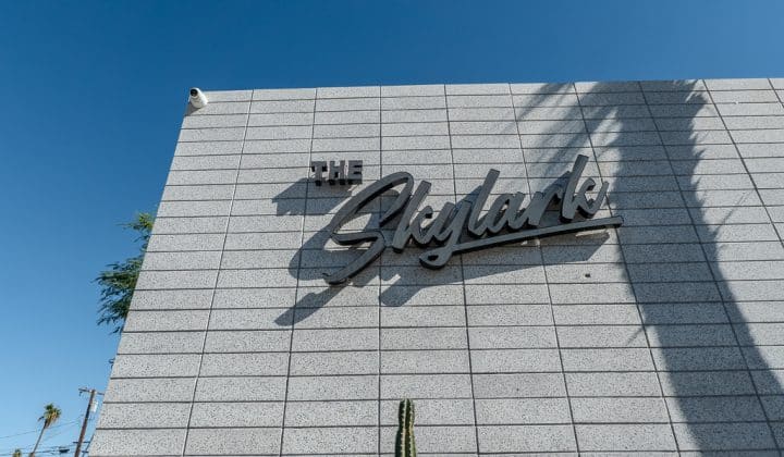 The image shows a building exterior with the sign "The Skylark" on a tiled wall. A security camera is mounted on the top left corner. A cactus is visible below. Sky is clear and blue.