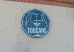 Circular sign on a wall features two toucans perched on a martini glass with the word "TOUCANS" below them.