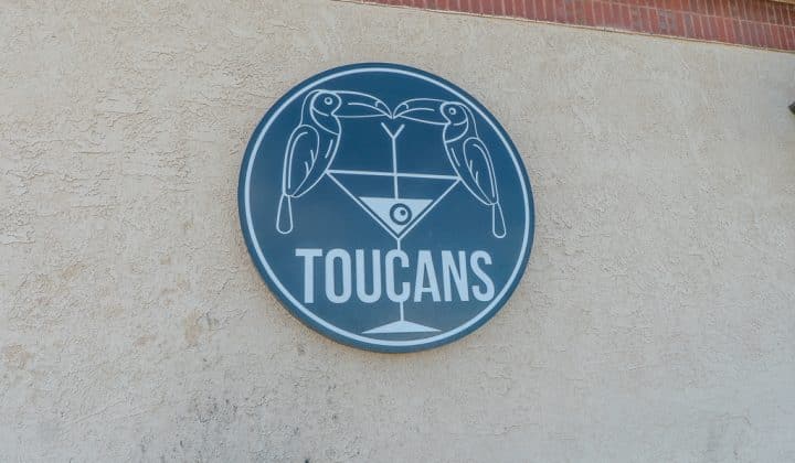 Circular sign on a wall features two toucans perched on a martini glass with the word "TOUCANS" below them.