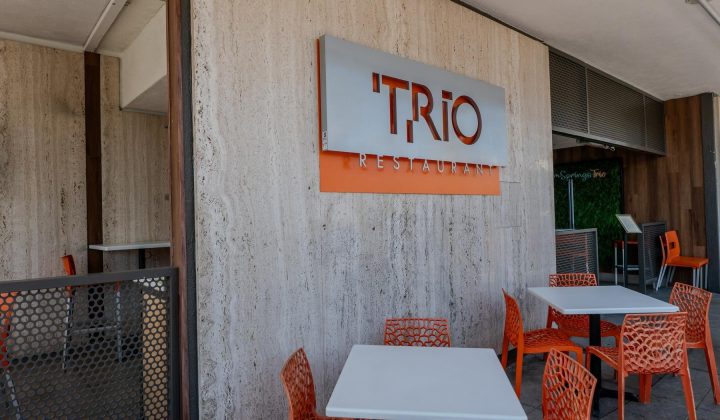 Outdoor seating of a restaurant with orange chairs and white tables. Sign reads "Trio Restaurant" on a wall.