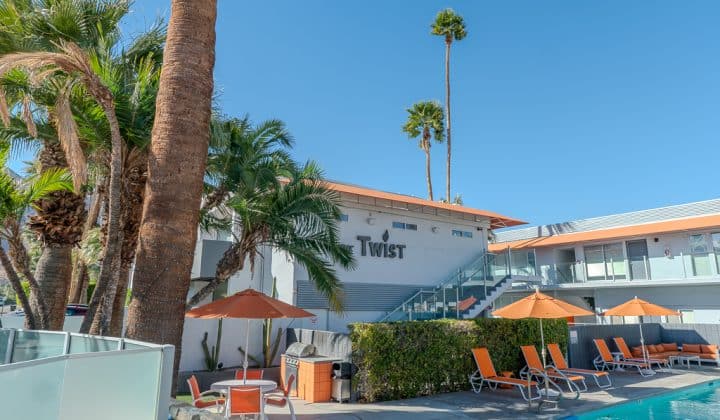 Poolside view of a modern hotel with orange lounge chairs and umbrellas, surrounded by palm trees under a clear blue sky.