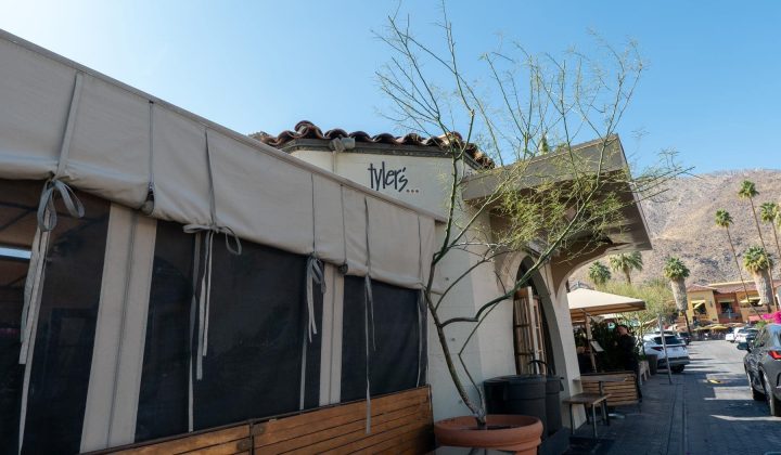 A building with beige awnings and a potted tree in front. Graffiti with "tykes..." is on the wall. Nearby are parked cars and a mountainous backdrop.