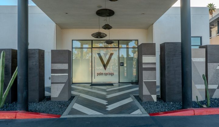 Entrance to a building with "V Palm Springs" on the glass door, surrounded by decorative pillars and cacti, viewed from the road.
