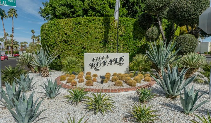 Sign reading "Villa Royale" surrounded by desert landscaping with cacti and succulents, against a backdrop of trees and a road sign.